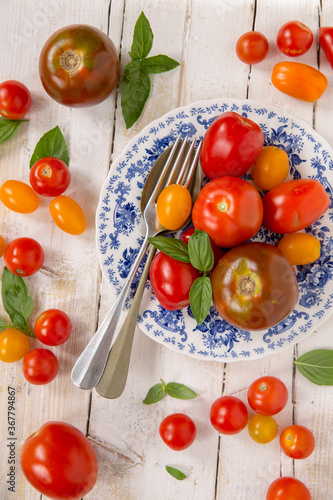 Piatto di pomodori di varietà diverse preso dall'alto su tavolo di legno bianco e pomodorini rossi e gialli con  foglie di basilico  photo