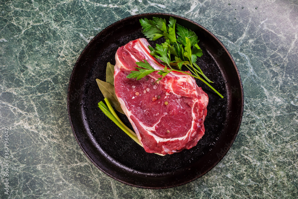 Fresh Raw Meat for Steak Ribeye, Decorated with Herbs and Sprinkled with Spices and Coarse Salt, Lies in a Cast-Iron Vintage Pan. View from Above. Green Marble Table. Fresh Raw Meat for Steak Ribeye, Decorated with Herbs and Sprinkled with Spices and Coarse Salt, Lies in a Cast-Iron Vintage Pan. View from Above. Green Marble Table.