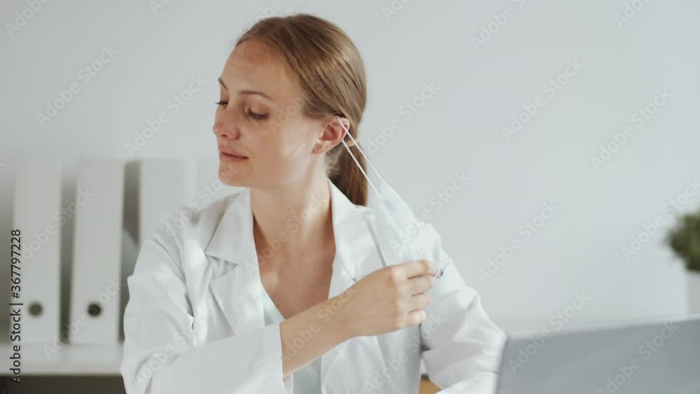 Tilt up shot of beautiful female doctor in lab coat with stethoscope over her neck removing medical face mask, smiling and looking at camera while sitting at desk in clinic Tilt up shot of beautiful female doctor in lab coat with stethoscope over her neck removing medical face mask, smiling and looking at camera while sitting at desk in clinic