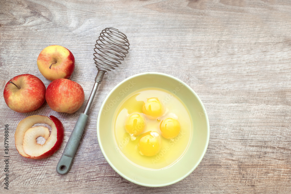 Ingridients for the pie: cutted sliced red apples and peel, broken eggs in plate, whisk on wooden table. Fresh ripe fruit background with copy space for text. Cooking concept. Ingridients for the pie: cutted sliced red apples and peel, broken eggs in plate, whisk on wooden table. Fresh ripe fruit background with copy space for text. Cooking concept.