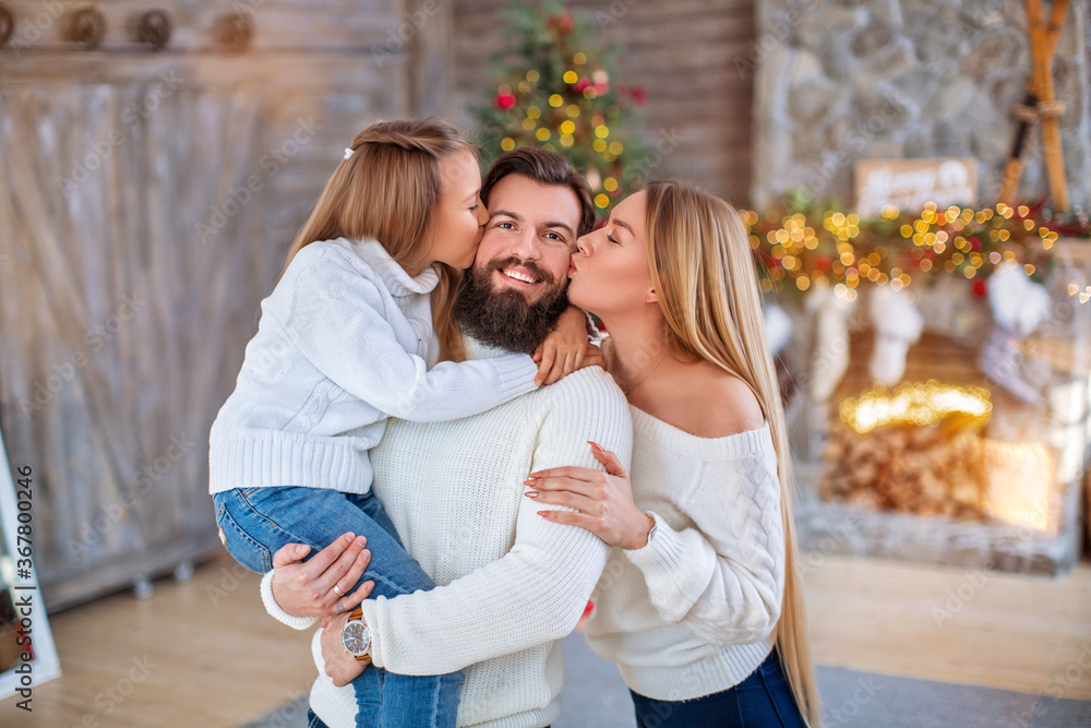 Fototapeta premium Family Christmas concept. Lovely family in bright clothes standing near decorated Xmas tree on winter evening. Happy household with daughter on floor at home near fireplace