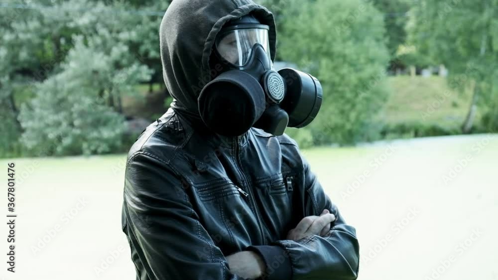 man in gas mask, black leather jacket with hood standing outdoors at forest near lake. protection from chemical weapons, virus epidemic. Post apocalyptic world. survivor, stalker after nuclear war man in gas mask, black leather jacket with hood standing outdoors at forest near lake. protection from chemical weapons, virus epidemic. Post apocalyptic world. survivor, stalker after nuclear war