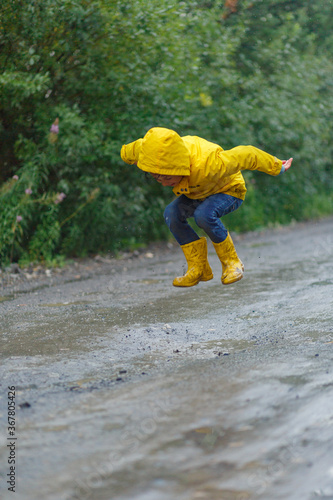 Kid playing in the rain in autumn park. Child jumping in muddy puddle on rainy fall day. Little boy in rain boots and yellow jacket outdoors in heavy shower. Kids waterproof footwear and coat. 