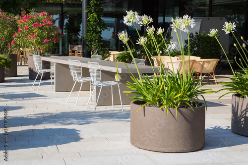 Moderne Garten und Terrassengestaltung: Hotel Restaurant Terrasse mit einem Tisch aus Stein und Stühlen aus Metall sowie Korbsessel und Regiestühle und weisse Schmucklilien in runden Pflanzgefässen