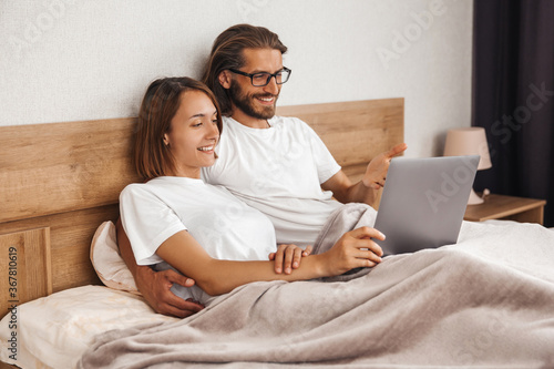 A young married couple is lying in bed at home and watching a movie on a laptop