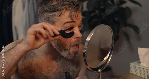 Handsome mature man applying mascara with opened mouth while sitting in front of mirror.Bearded guy with eye patches getting ready for date in evening. Home background.