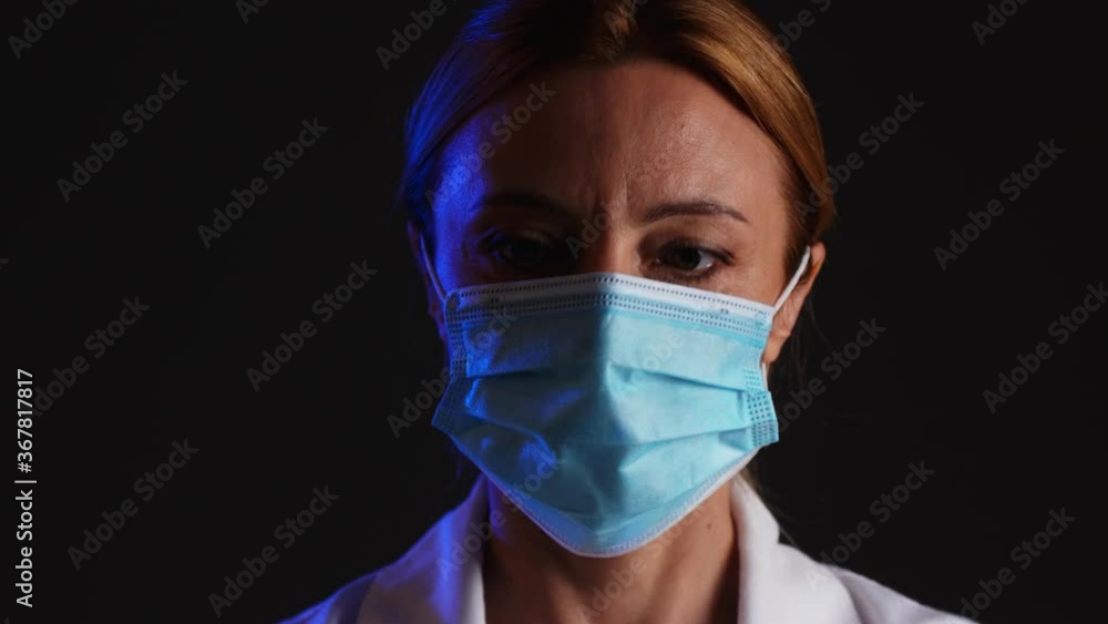 Mature female doctor putting on protective medical mask, hat and eyeglasses against dark background Mature female doctor putting on protective medical mask, hat and eyeglasses against dark background