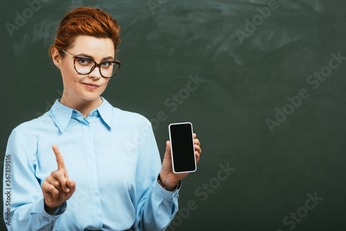 sceptical teacher showing forbidding gesture while holding smartphone with blank screen near chalkboard