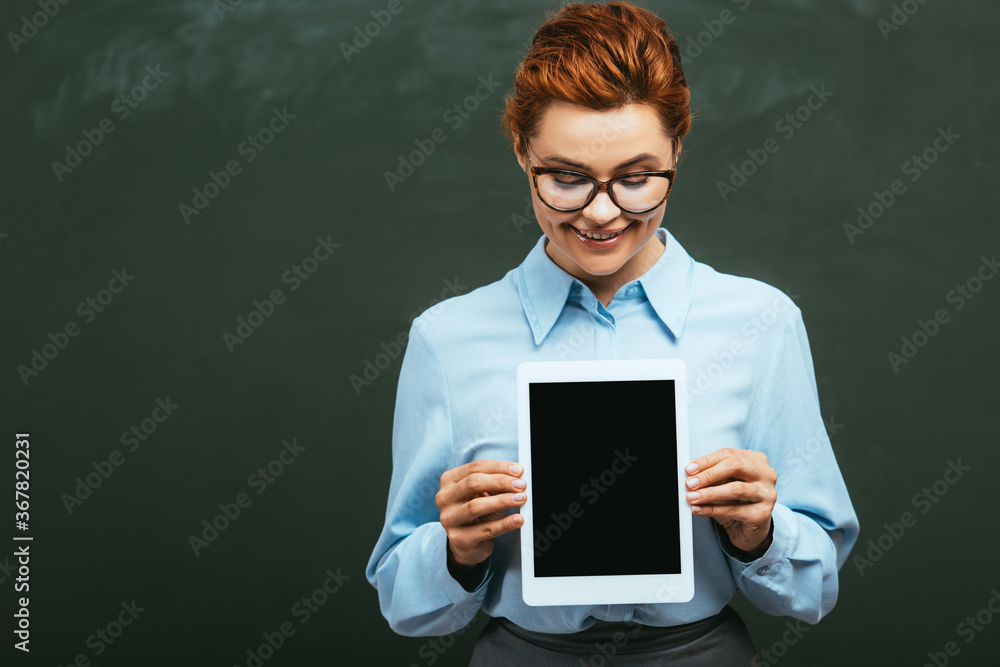 smiling teacher holding digital tablet with blank screen while standing near chalkboard smiling teacher holding digital tablet with blank screen while standing near chalkboard
