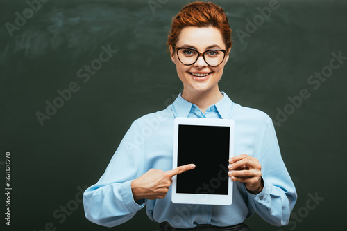 happy teacher pointing with finger at digital tablet with blank screen near chalkboard
