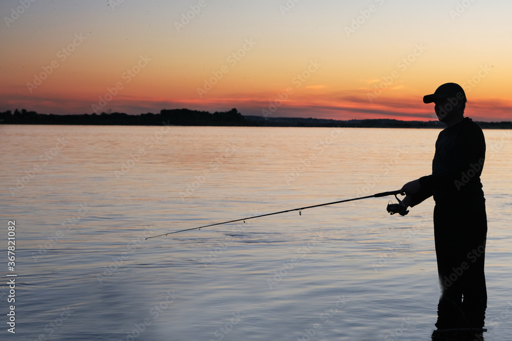 Fisher man fishing with spinning rod on a river bank at misty sunset