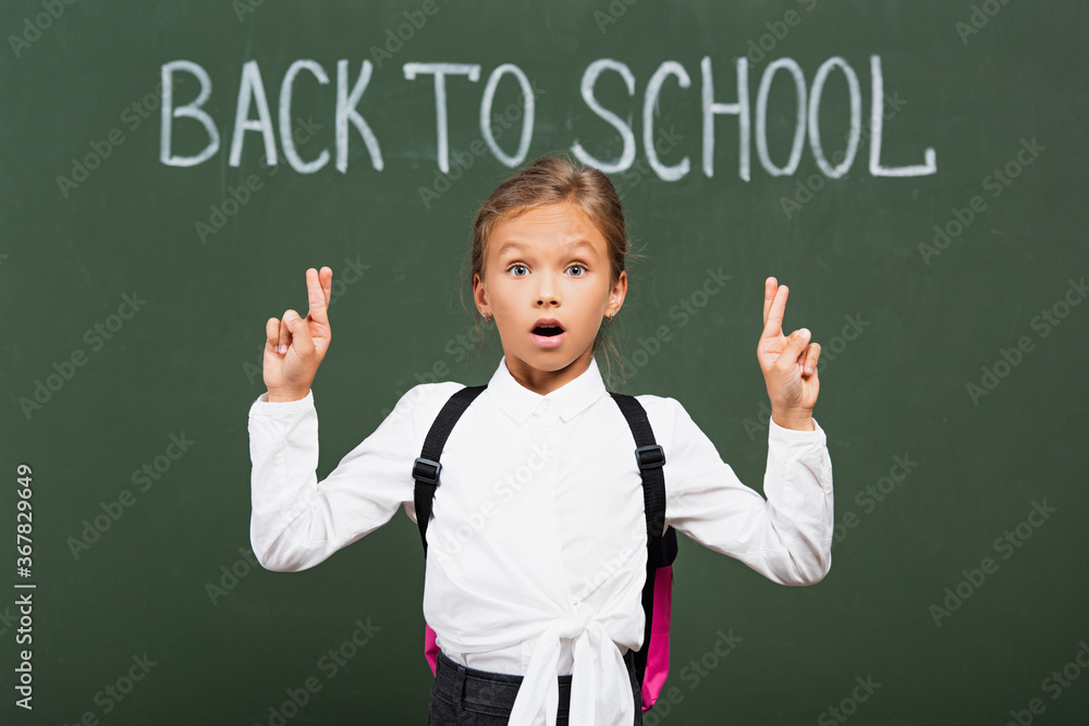 shocked schoolgirl holding crossed fingers near back to school inscription on chalkboard