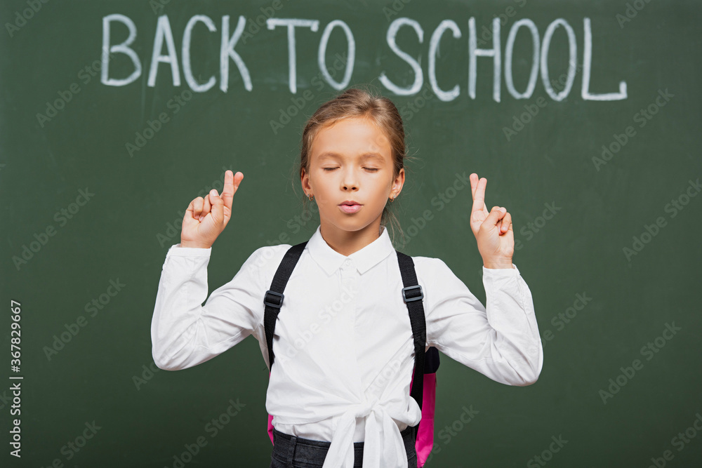 adorable schoolgirl with closed eyes holding crossed fingers near back to school inscription on chalkboard