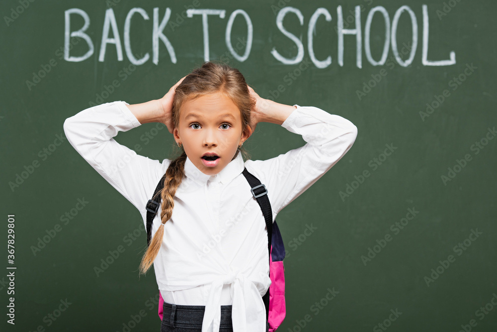 shocked schoolgirl touching head near chalkboard with back to school lettering