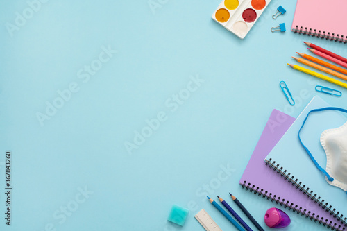 Education concept. Colorful stationery set on the blue background. Top view of blue, pink and purple notebooks, paper pins, eraser, felt tip pens, color pencils, watercolor and face mask. Copy space. 