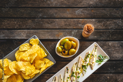 Three dishes with potato chips, picked anchovies and green olives on a dark wooden table with toothpicks. Typical Spanish snacks to eat in a bar. 