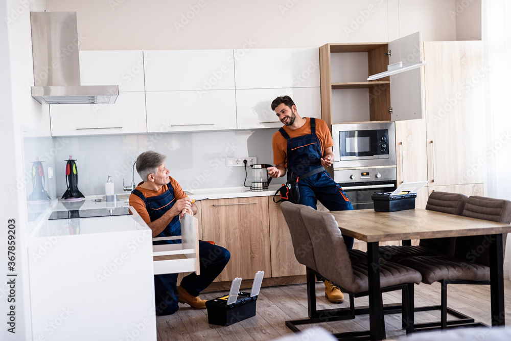 Obraz premium Full length shot of two handymen, workers in uniform talking while assembling kitchen cabinet using screwdriver indoors. Furniture repair and assembly concept