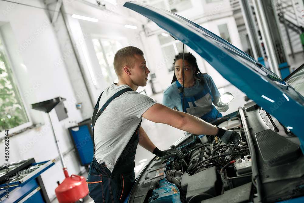 Obraz premium Apprenticeship. Professional male mechanic repairing car engine, tighten, screw with spanner while his female colleague looking at him, holding torch under car hood at service station