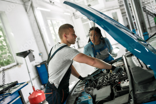 Apprenticeship. Professional male mechanic repairing car engine, tighten, screw with spanner while his female colleague looking at him, holding torch under car hood at service station