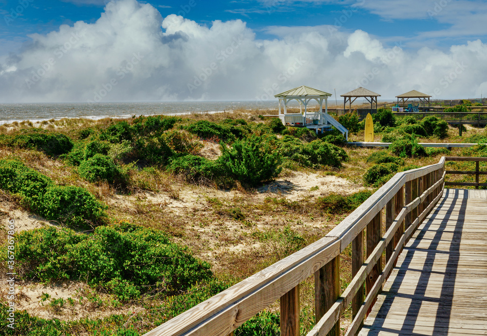 A beach boardwalk over sand dunes with dramatic clouds and gazebos in the background in HDR.