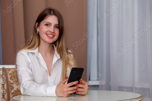 Girl of Slavic appearance with phone and white Cup is sitting at table. Female takes actor's emotions. Young charming woman is sitting in hotel room and drinking coffee or tea with smartphone