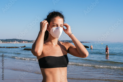 Young girl in a bikini with a mask. Wearing the coronavirus mask on the beach. Wearing the medical face mask in public places. Coronavirus quarantine. Vacation and travel in the Covid-19 pandemic.