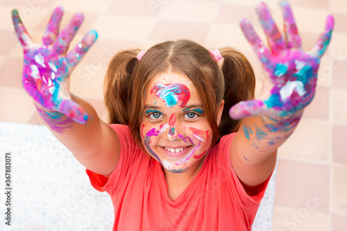 The happy girl was smeared with paint.  A child with bright colors on his face  hands and hair smiles and shows smiles on his hands
