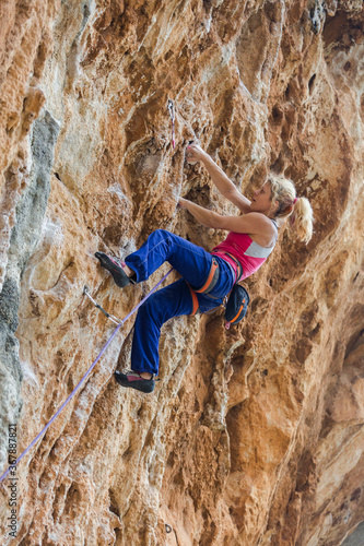 Side view of the young woman rock climber in bright blue pants climbing on the vertical challenging rock wall