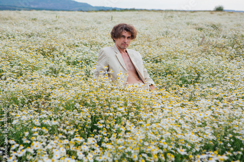 Tall handsome man sitting on a white chair in camomile flowers field