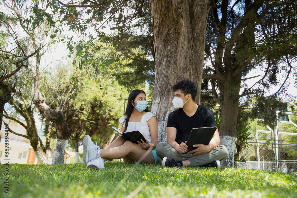 Young Hispanic couple in the park with disposable face masks - young people studying with an electronic tablet and a book in a natural park - social distancing - general shot Young Hispanic couple in the park with disposable face masks - young people studying with an electronic tablet and a book in a natural park - social distancing - general shot