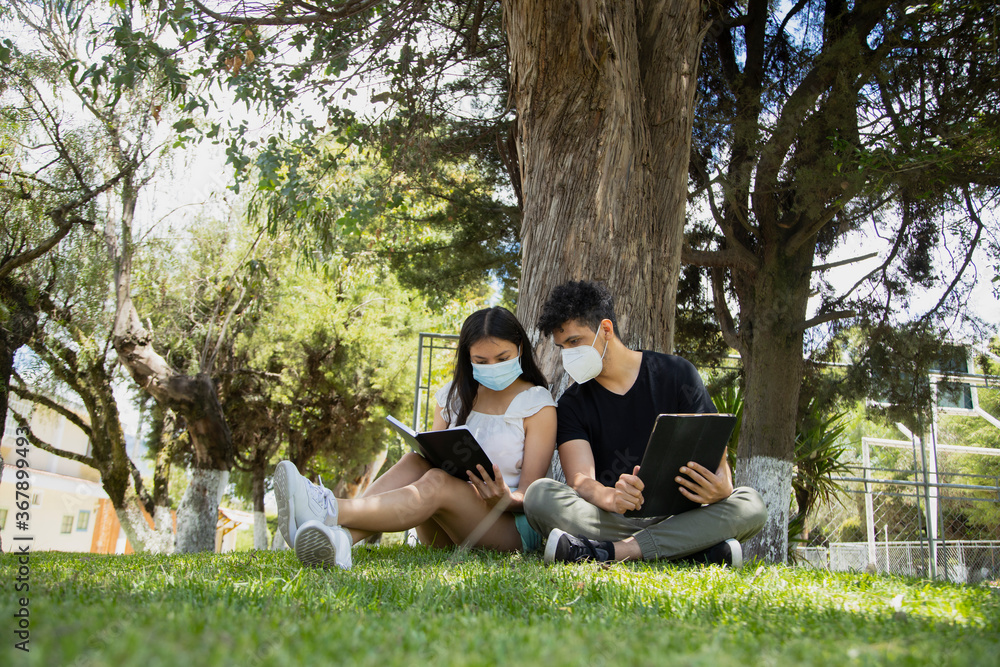Young Hispanic couple in the park with disposable face masks - young people studying with an electronic tablet and a book in a natural park - social distancing - general shot Young Hispanic couple in the park with disposable face masks - young people studying with an electronic tablet and a book in a natural park - social distancing - general shot