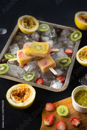 Homemade tropical ice cream or popsicles garnished with pieces of ice, passion fruit, kiwi and strawberries with leaves, top view