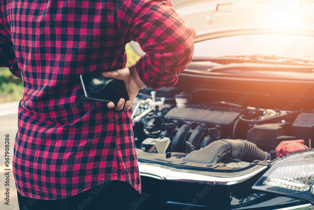 Fototapeta premium Young woman using mobile phone while looking at broken down car on road.