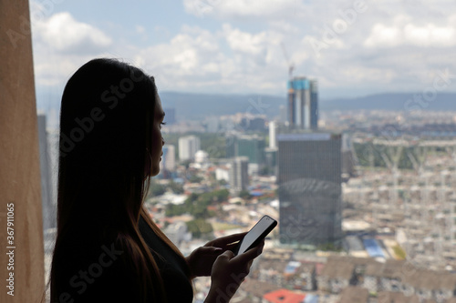 Young beautiful modern Malay woman using smart phone video chat next to window cityscape cloudy blue sky