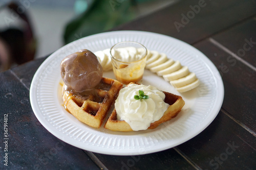 A plate of Belgian waffle with banana and caramel sauce, topped with whipped cream and chocolate ice cream.