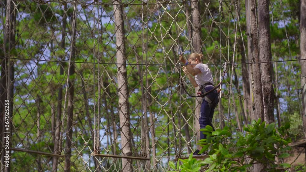 A young woman in an adventure park. She wears a safety harness. She climbs on a high rope trail. Outdoor amusement center with climbing activities consisting of zip lines and all sorts of obstacles A young woman in an adventure park. She wears a safety harness. She climbs on a high rope trail. Outdoor amusement center with climbing activities consisting of zip lines and all sorts of obstacles
