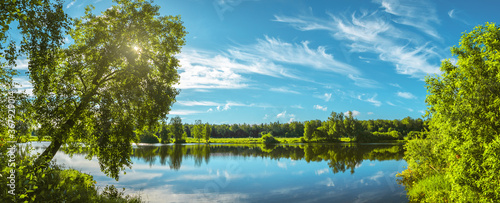 Beautiful summer sunrise nature landscape. Tranquil morning scene with warm sunlight and amazing reflection of clods in sky on the water surface. Calm lake and green trees at sunrise.Panorama.