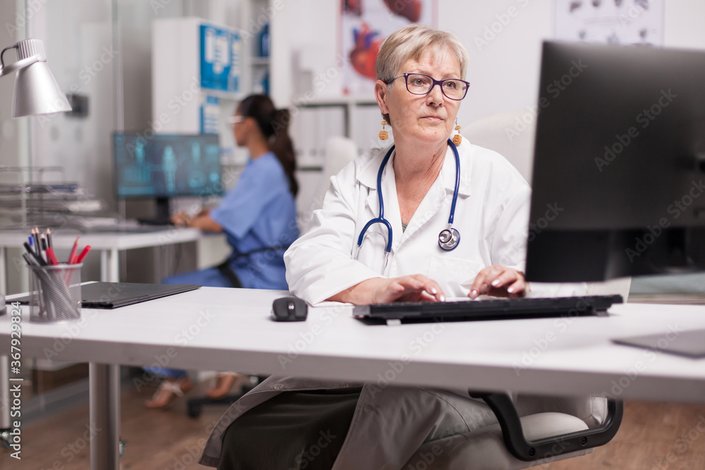Experienced doctor using computer in clinic cabinet and nurse typing on pc in the background. Experienced doctor using computer in clinic cabinet and nurse typing on pc in the background.