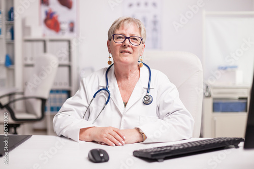 Senior medical practitioner with stethoscope in hospital cabinet smiling to camera.