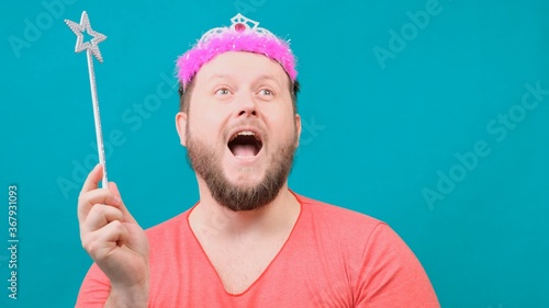 funny bearded freaky man in a pink T-shirt with a deadema on his head is dreaming with a magic wand in his hand. A funny wizard joke to make and fulfill a wish.
