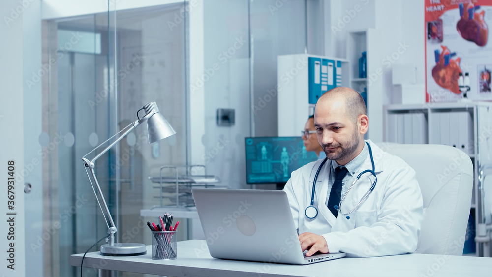 Doctor typing an e-mail from modern office of private clinic with glass walls, nurse in background checking X Rays, medical staff and patients walking in busy hallway. Healthcare system clinic Doctor typing an e-mail from modern office of private clinic with glass walls, nurse in background checking X Rays, medical staff and patients walking in busy hallway. Healthcare system clinic
