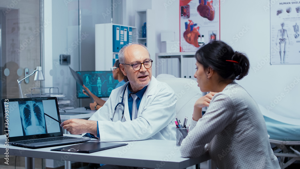 Obraz premium Doctor analyzing X ray scan results on laptop with young female patient. Elderly senior experienced doctor talking with patient about lungs, x ray pneumonia, cancer, examination specialist consulting