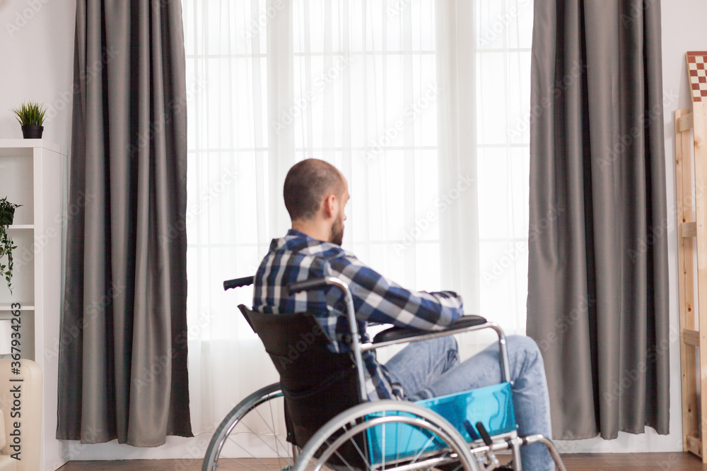 Disabled person sits in wheelchair against window. Serious sad caucasian man wearing casual clothes and look at large panoramic view in bright modern living room, hospital or clinic. Disabled person sits in wheelchair against window. Serious sad caucasian man wearing casual clothes and look at large panoramic view in bright modern living room, hospital or clinic.