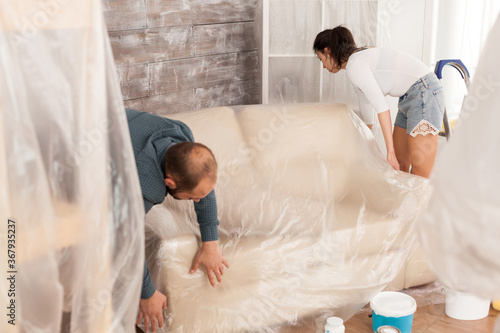 Couple wrapping sofa in plastic foil for protection while they are renovate living room. Home during renovation, decoration and painting. Interior apartment improvement maintenance. Roller, ladder for