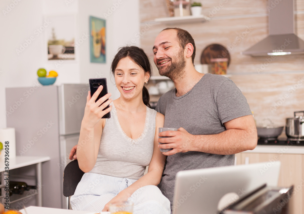 Cheerful couple talking with friends on video call in the morning sitting on the chair in the kitchen. Joyful married husband and wife making funny faces while taking a photo during breakfast in Cheerful couple talking with friends on video call in the morning sitting on the chair in the kitchen. Joyful married husband and wife making funny faces while taking a photo during breakfast in