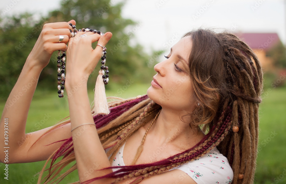 Young beautiful girl with long dreadlocks holds in hands the necklace made by natural stones for meditation outdoors during morning on green grass of her backyard