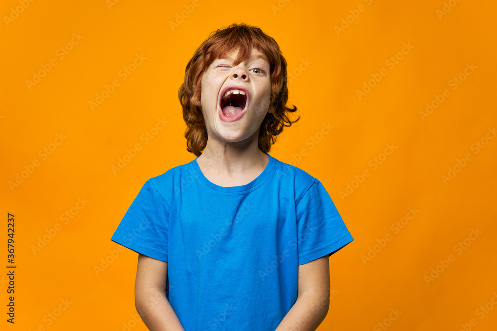 An emotional child with a wide open mouth yawns blue t-shirt yellow background red hair An emotional child with a wide open mouth yawns blue t-shirt yellow background red hair