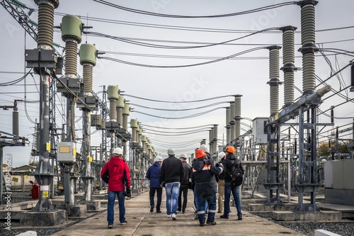 Backs of engineers and journalists with cameras exploring modern electric power station and looking at high voltage power lines and transmission towers.