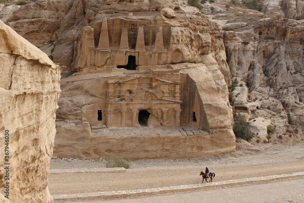 Obelisk tomb in ancient Petra city in Jordan. The building is carved out of sandstone right into the rock. Unrecognisable horseman rides on the road. Theme of travel in Jordan.