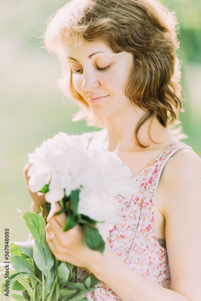 Naklejka premium Beautiful young Caucasian blond woman in light dress holding bouquet of white peonies, walking in summer field or garden in sunset. Woman with flowers outdoors